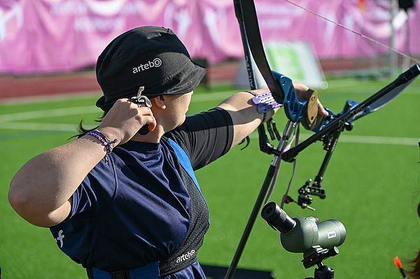 Eine Sch&uuml;tzin zielt mit einem Bogen auf ein Ziel, w&auml;hrend sie auf einer gr&uuml;nen Sportfl&auml;che steht.