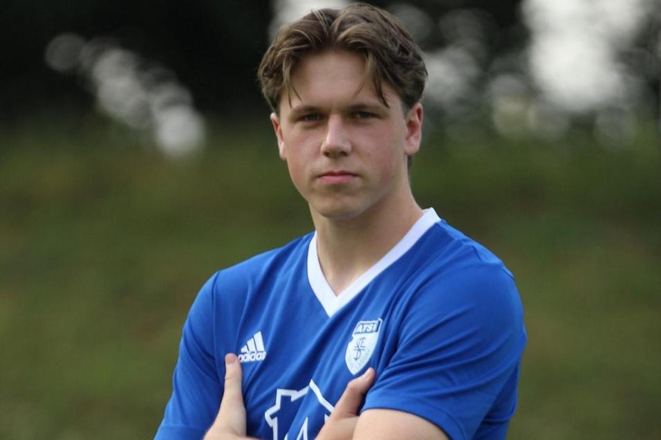 Ein junger Mann im blauen Fußballtrikot posiert mit verschränkten Armen auf einem grünen Sportplatz.