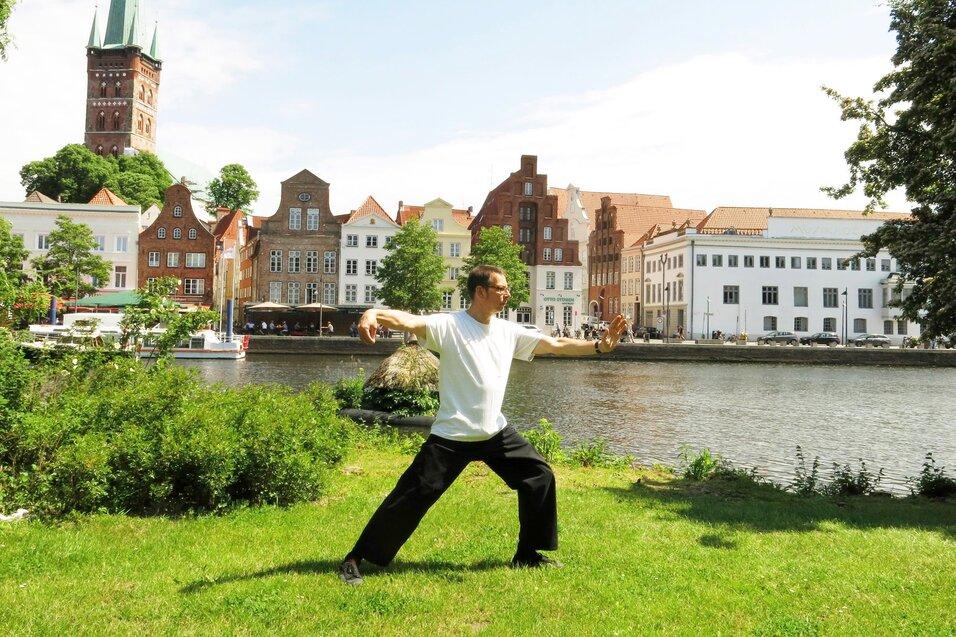Mann in Rückenansicht praktiziert Tai Chi auf einer Wiese am Ufer eines Flusses, historische Gebäude im Hintergrund.