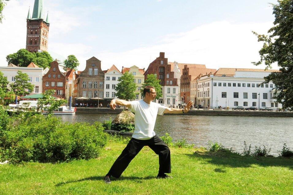 Mann in Rückenansicht praktiziert Tai Chi auf einer Wiese am Ufer eines Flusses, historische Gebäude im Hintergrund.