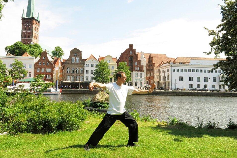 Mann in Rückenansicht praktiziert Tai Chi auf einer Wiese am Ufer eines Flusses, historische Gebäude im Hintergrund.