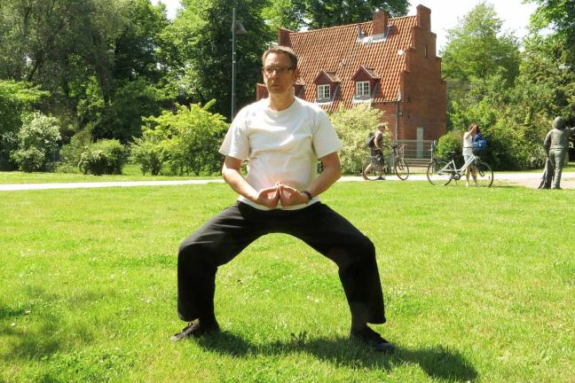 Mann in heller Kleidung steht in einer meditierenden Position auf einer grünen Wiese; im Hintergrund Fahrräder und Personen.