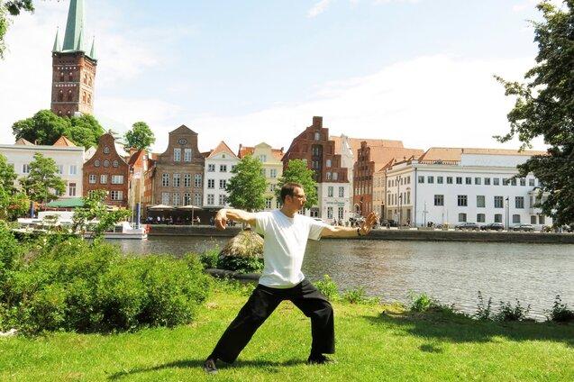 Mann in R&uuml;ckenansicht praktiziert Tai Chi auf einer Wiese am Ufer eines Flusses, historische Geb&auml;ude im Hintergrund.