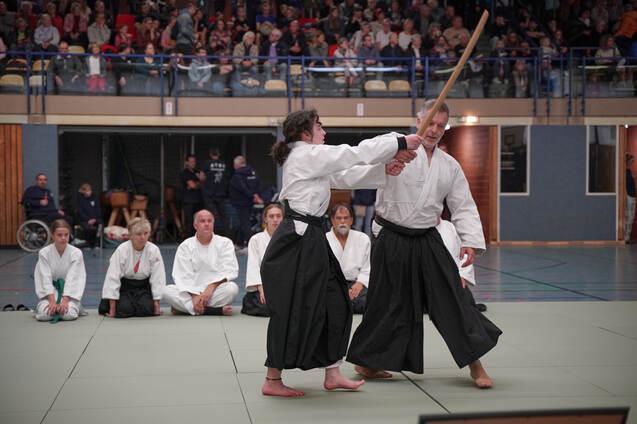 Zwei Aikidoka in wei&szlig;er &Uuml;bungskleidung demonstrieren eine Technik mit Holzschwert vor einer Zuschauergruppe.