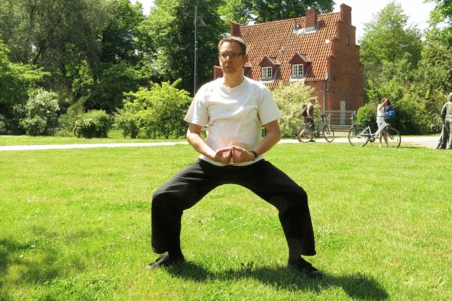 Mann in heller Kleidung steht in einer meditierenden Position auf einer grünen Wiese; im Hintergrund Fahrräder und Personen.