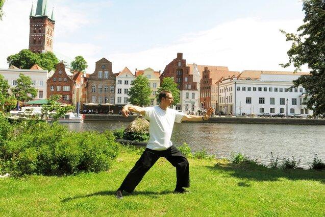 Mann in R&uuml;ckenansicht praktiziert Tai Chi auf einer Wiese am Ufer eines Flusses, historische Geb&auml;ude im Hintergrund.