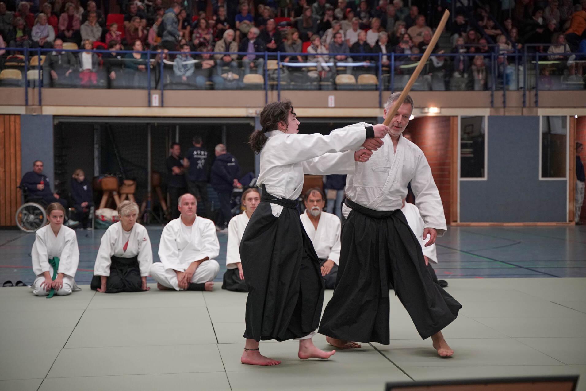 Zwei Aikidoka in weißer Übungskleidung demonstrieren eine Technik mit Holzschwert vor einer Zuschauergruppe.