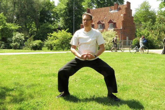 Mann in heller Kleidung steht in einer meditierenden Position auf einer grünen Wiese; im Hintergrund Fahrräder und Personen.