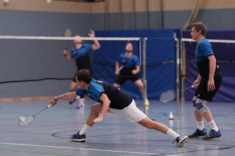 Zwei Badmintonspieler in blauen Shirts und Shorts; ein Spieler greift den Ball während eines Matches in einer Halle.