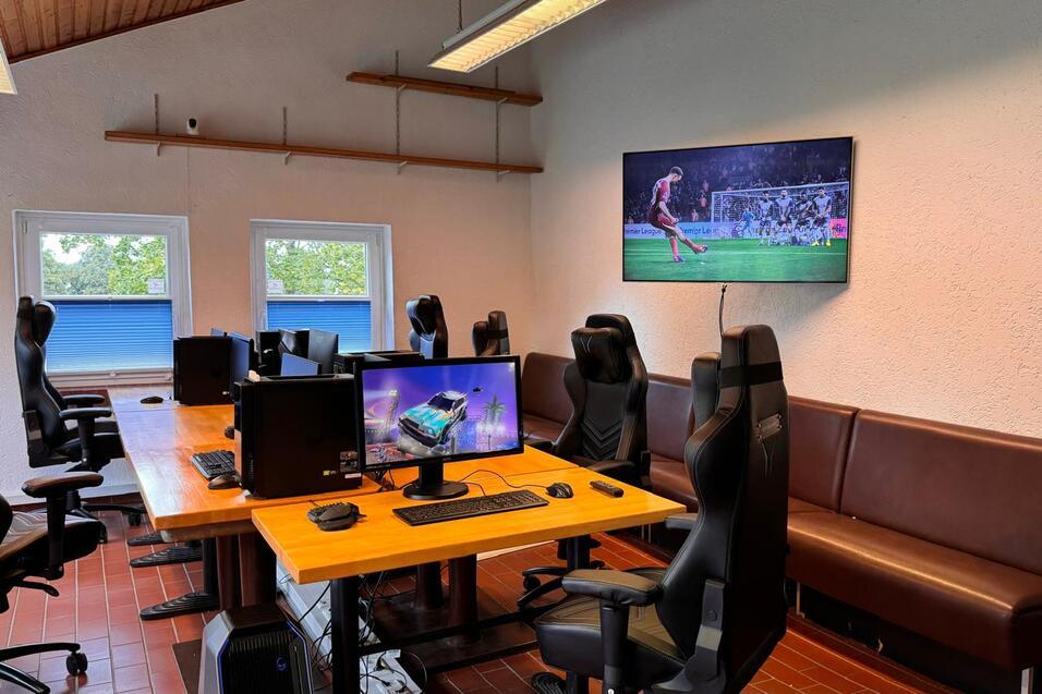 B&uuml;ro mit mehreren Gaming-PCs und ergonomischen St&uuml;hlen, gro&szlig;er Bildschirm zeigt ein Fu&szlig;ballspiel.