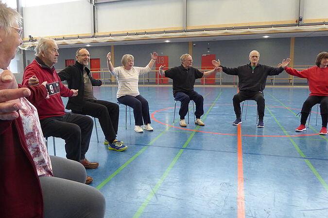 Gruppe &auml;lterer Menschen sitzt auf St&uuml;hlen in einer Sporthalle und f&uuml;hrt armbewegende &Uuml;bungen durch.