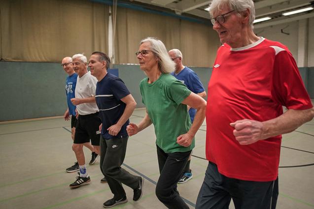 Sechs Erwachsene joggen in Sportkleidung in einer Turnhalle auf einem grauen Boden.