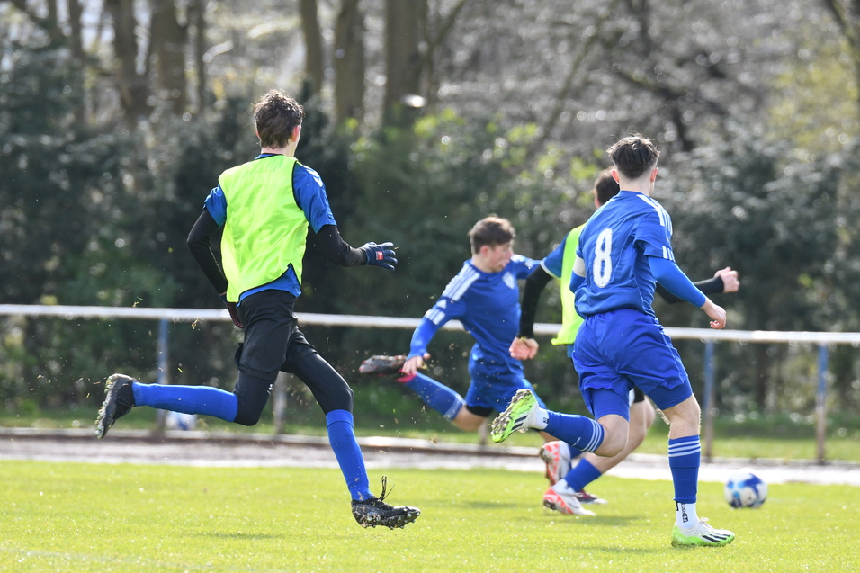 Vier Fu&szlig;ballspieler in blauen Trikots laufen auf einem Rasenplatz, ein Spieler tr&auml;gt ein gr&uuml;nes Trikot.