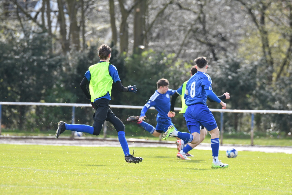 Vier Fu&szlig;ballspieler in blauen Trikots laufen auf einem Rasenplatz, ein Spieler tr&auml;gt ein gr&uuml;nes Trikot.