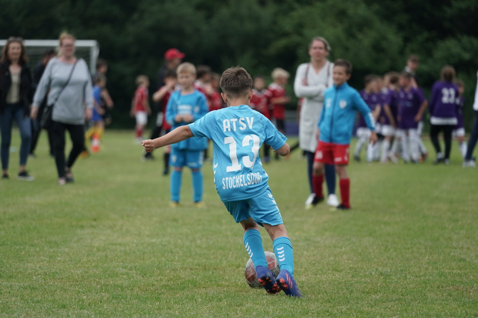 Junge in blauer Sportkleidung mit der Nummer 13 dribbelt einen Fu&szlig;ball auf einem Rasenplatz, Zuschauer im Hintergrund.