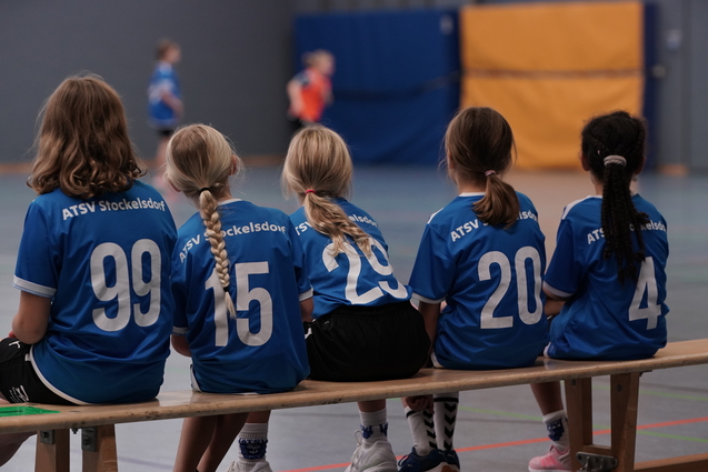 F&uuml;nf Kinder in blauen Sporttrikots sitzen auf einer Bank in einer Sporthalle, R&uuml;cken zur Kamera.