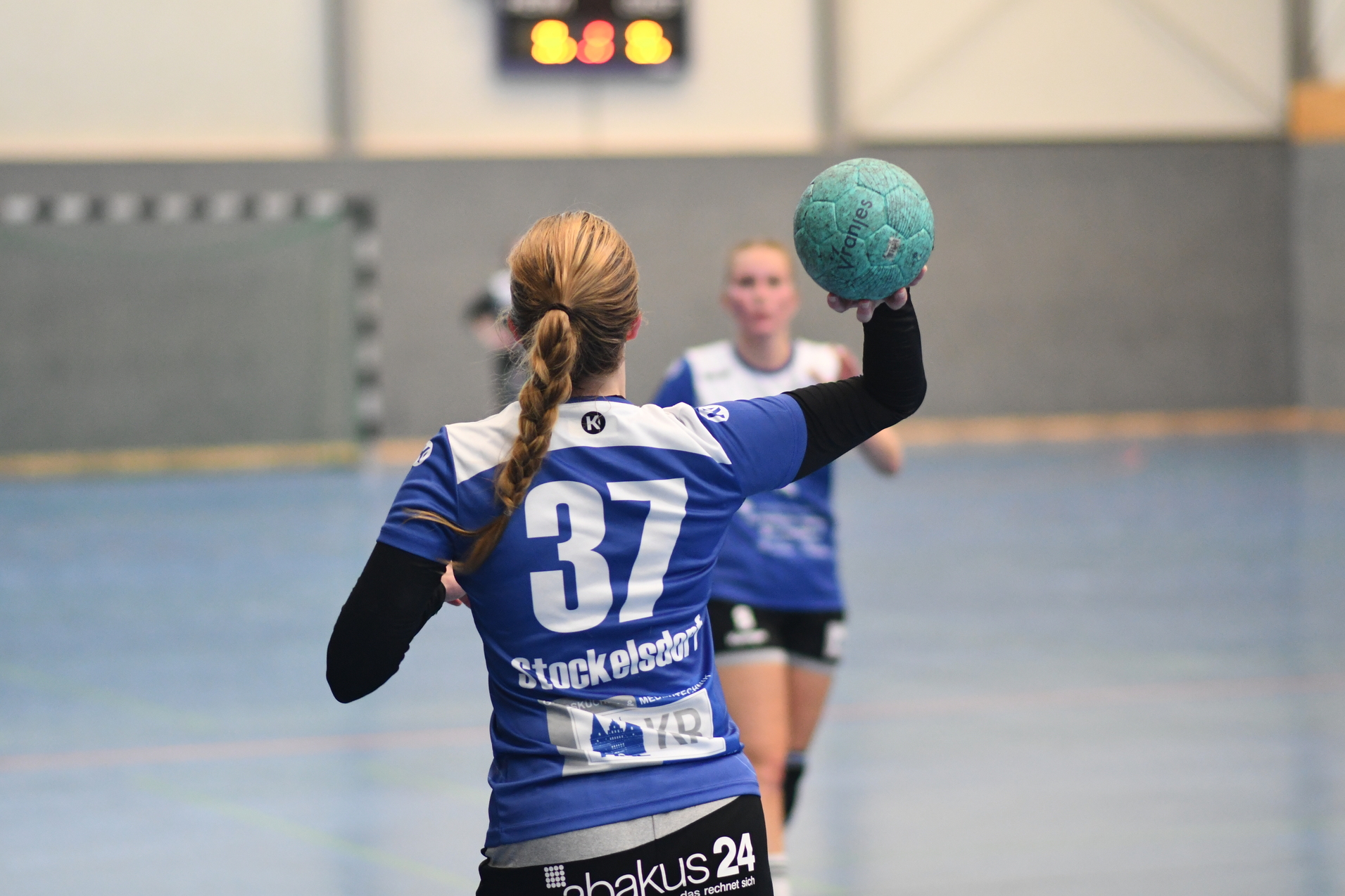 Handballspielerin mit der Nummer 37 hebt einen blauen Ball zur Abgabe eines Abwurfs in einer Sporthalle.