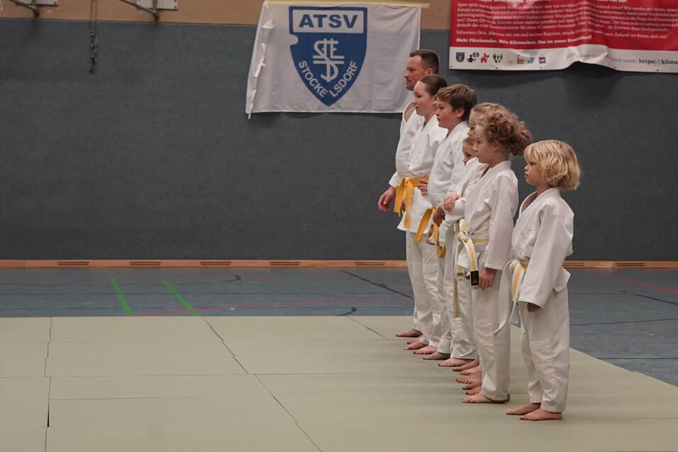 Gruppe von Judokas in weißen Anzügen mit gelben Gurten steht auf einer Matte in der Sporthalle, bereit für das Training.