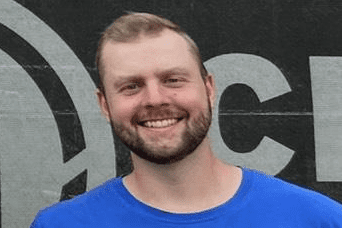 Lächelnder Mann in einem blauen T-Shirt mit Logo vor einer grauen Wand mit grafischen Elementen.