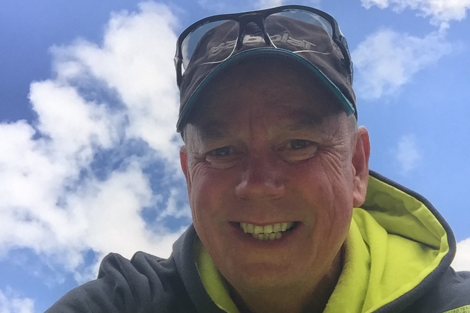 Mann mit Sonnenbrille l&auml;chelt, w&auml;hrend er eine Kiste mit gelben Tennisb&auml;llen vor blauem Himmel h&auml;lt.