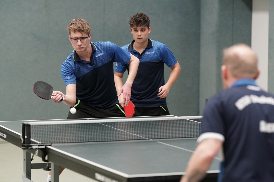 Zwei Spieler in blauen T-Shirts stehen an einem Tischtennistisch, bereit, einen Ball zu spielen.