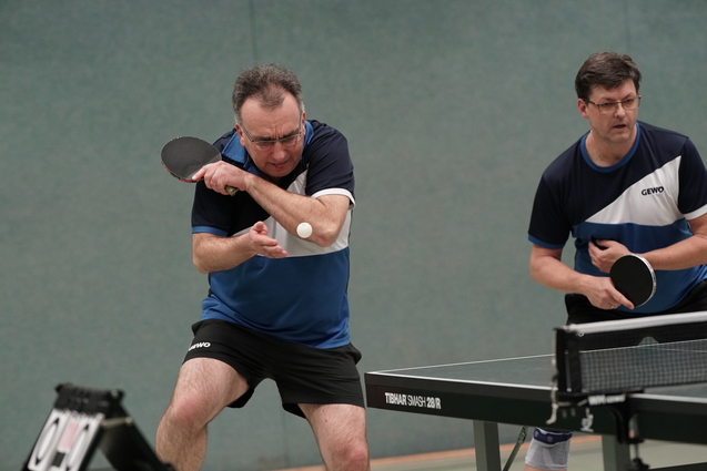 Zwei M&auml;nner spielen Tischtennis in Sportkleidung, einer schl&auml;gt den Ball mit einem Schl&auml;ger auf einem Tisch.