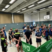 Gruppe von Tischtennisspielern h&ouml;rt einem Trainer in einer Sporthalle aufmerksam zu. Tischtennistische sind im Hintergrund sichtbar.