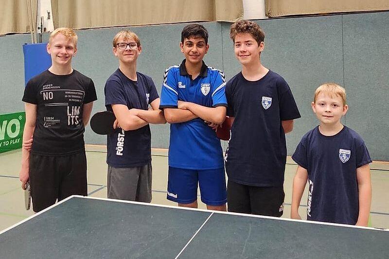 F&uuml;nf Jugendliche stehen l&auml;chelnd vor einem Tischtennistisch in einer Sporthalle.