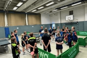 Gruppe von Tischtennisspielern h&ouml;rt einem Trainer in einer Sporthalle aufmerksam zu. Tischtennistische sind im Hintergrund sichtbar.