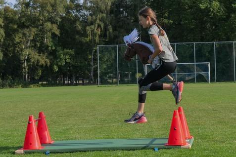 Mädchen springt mit einem Steckenpferd über zwei orangefarbene Pylonen auf einer Wiese. Hintergrund mit Fußballtoren.