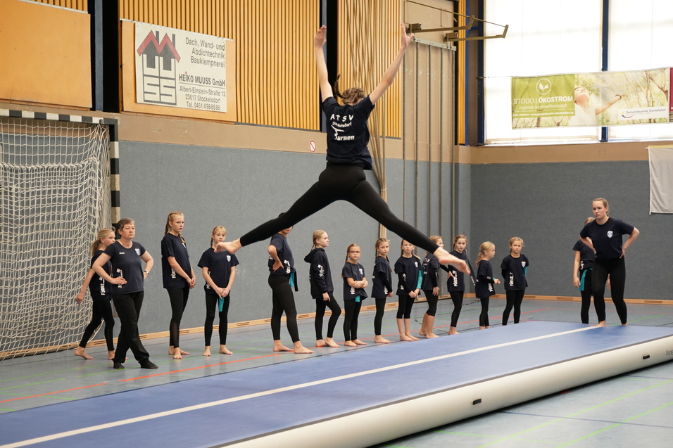 Eine Turnerin springt agil von einem Trampolin, w&auml;hrend eine Gruppe M&auml;dchen auf der Seite zuschaut.