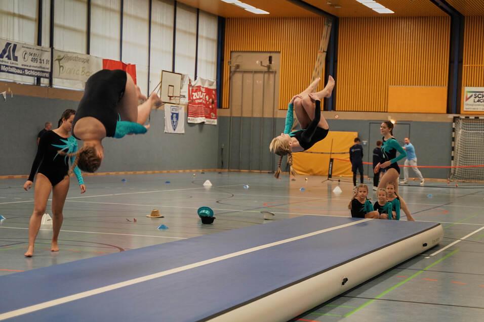 Zwei Turnerinnen führen saltoartige Sprünge auf einem Luftkissen in einer Sporthalle aus.
