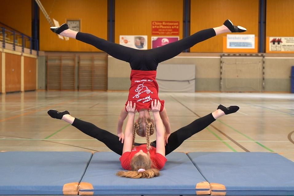 Zwei Akrobatinnen in roten T-Shirts führen eine Balanceübung auf einer Gymnastikmatte in einer Sporthalle aus.