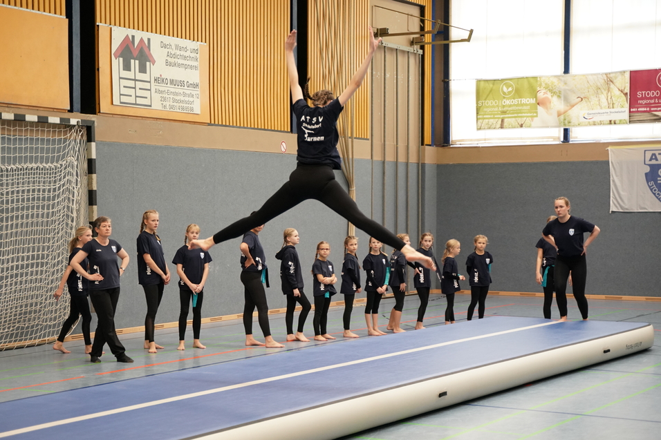 Eine Turnerin springt agil von einem Trampolin, w&auml;hrend eine Gruppe M&auml;dchen auf der Seite zuschaut.