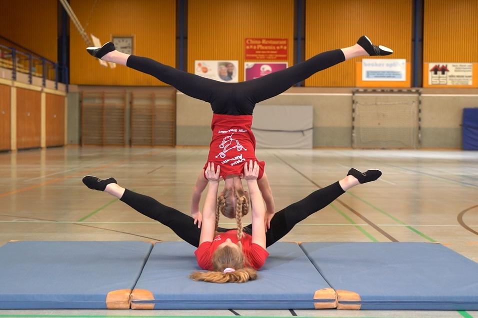 Zwei Akrobatinnen in roten T-Shirts f&uuml;hren eine Balance&uuml;bung auf einer Gymnastikmatte in einer Sporthalle aus.