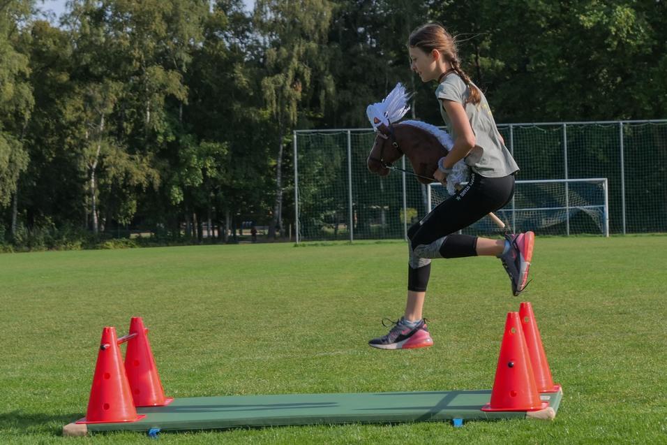 Mädchen springt mit einem Steckenpferd über zwei orangefarbene Pylonen auf einer Wiese. Hintergrund mit Fußballtoren.