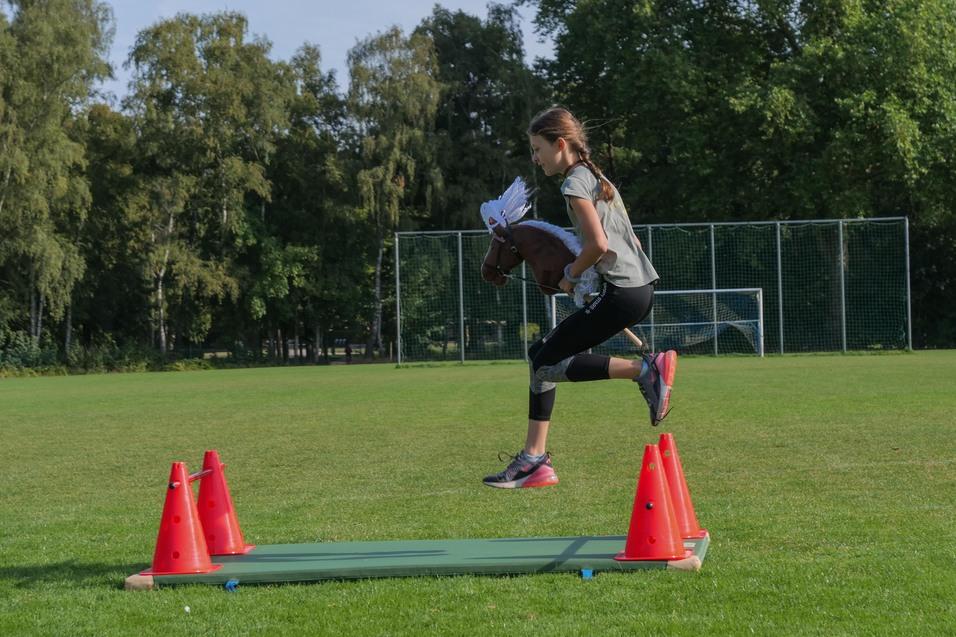 Mädchen springt mit einem Steckenpferd über zwei orangefarbene Pylonen auf einer Wiese. Hintergrund mit Fußballtoren.