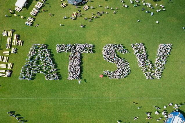Vogelperspektive auf Menschen, die das Wort "ATSV" auf einer Rasenfl&auml;che bilden, umgeben von Tischen und St&uuml;hlen.