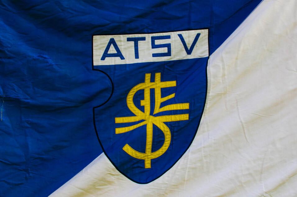 Fahne des ATSV in blau-wei&szlig; mit gelbem Wappen und Fransen, im Hintergrund Menschen auf einer Veranstaltung.