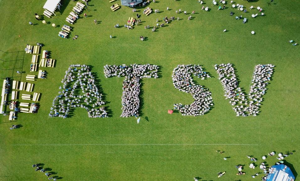 Vogelperspektive auf Menschen, die das Wort "ATSV" auf einer Rasenfläche bilden, umgeben von Tischen und Stühlen.