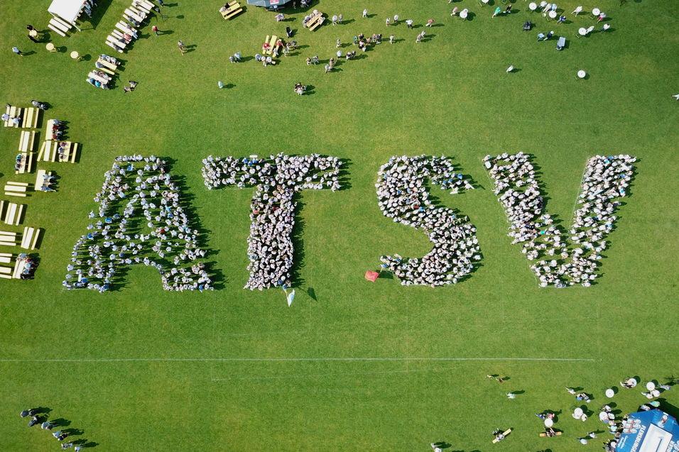Vogelperspektive auf Menschen, die das Wort "ATSV" auf einer Rasenfl&auml;che bilden, umgeben von Tischen und St&uuml;hlen.