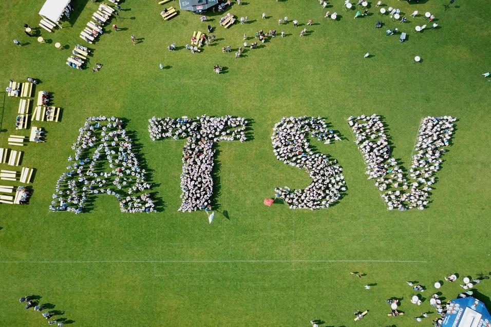 Vogelperspektive auf Menschen, die das Wort "ATSV" auf einer Rasenfl&auml;che bilden, umgeben von Tischen und St&uuml;hlen.