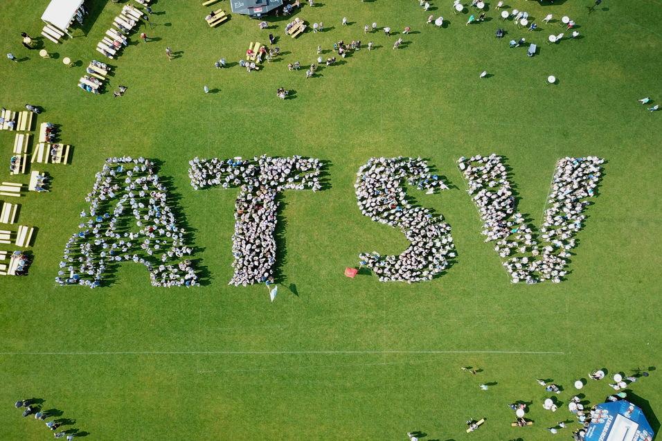 Vogelperspektive auf Menschen, die das Wort "ATSV" auf einer Rasenfl&auml;che bilden, umgeben von Tischen und St&uuml;hlen.