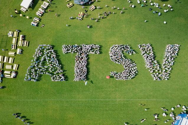 Vogelperspektive auf Menschen, die das Wort "ATSV" auf einer Rasenfl&auml;che bilden, umgeben von Tischen und St&uuml;hlen.