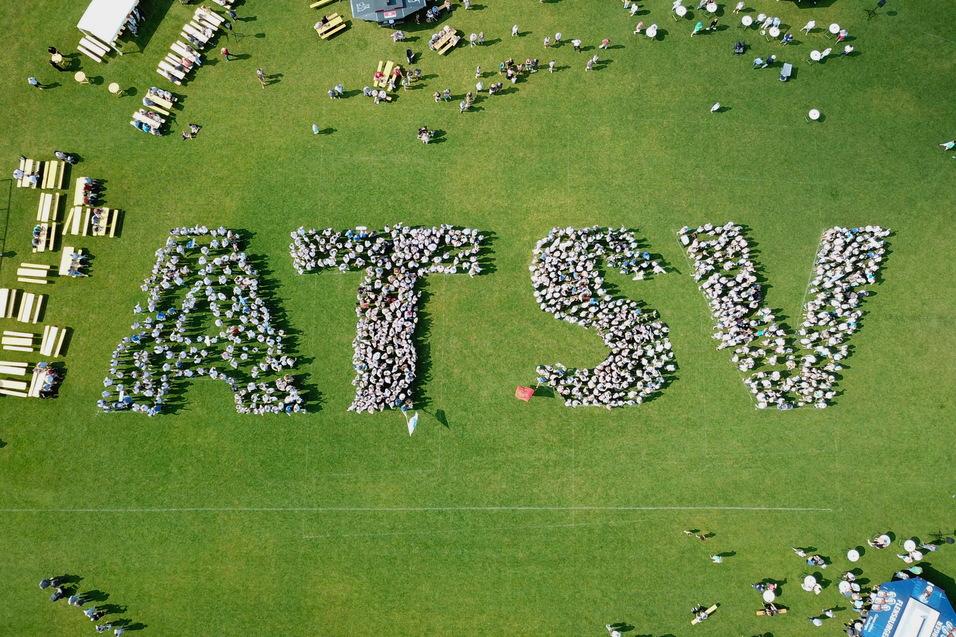 Vogelperspektive auf Menschen, die das Wort "ATSV" auf einer Rasenfl&auml;che bilden, umgeben von Tischen und St&uuml;hlen.