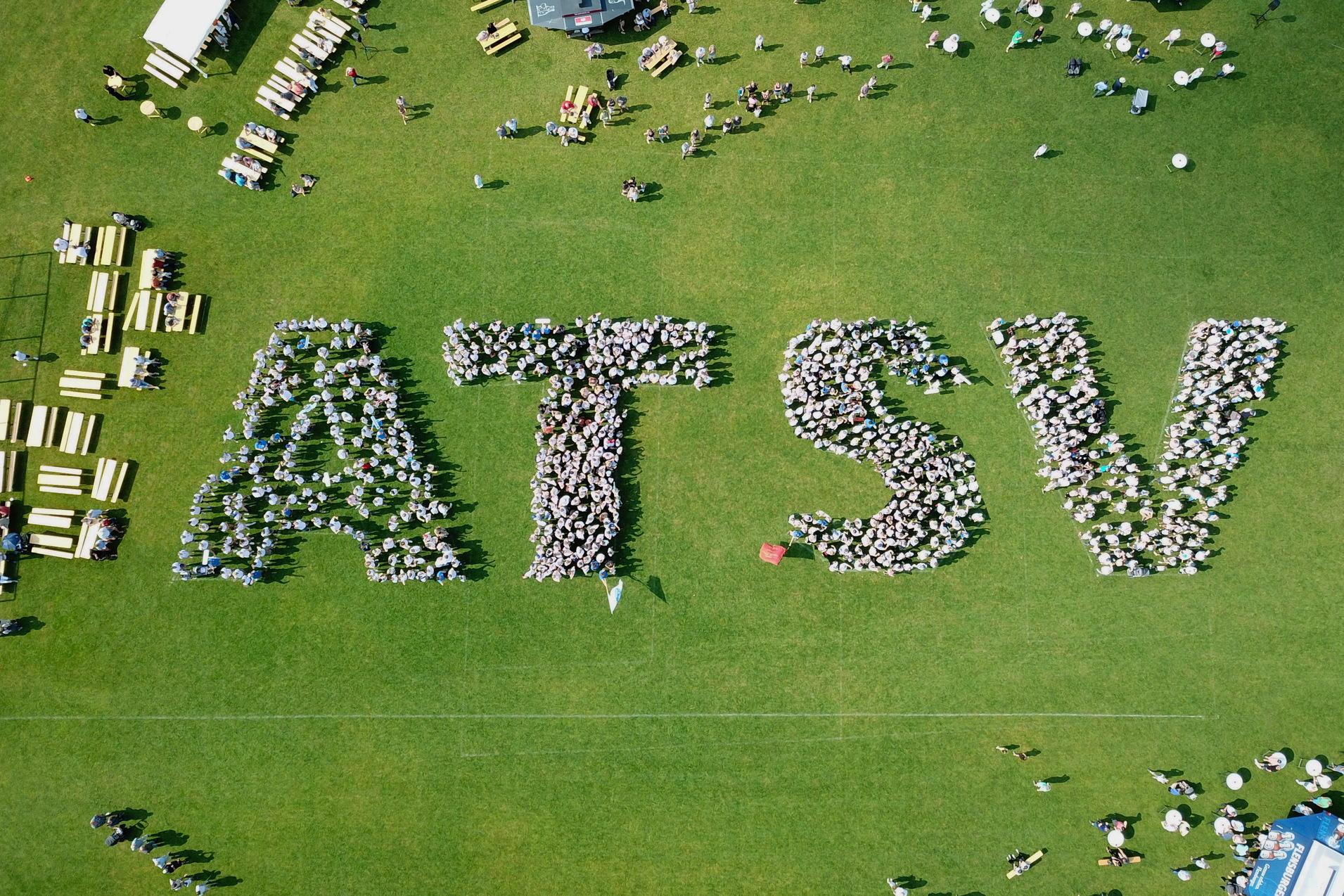 Vogelperspektive auf Menschen, die das Wort "ATSV" auf einer Rasenfl&auml;che bilden, umgeben von Tischen und St&uuml;hlen.