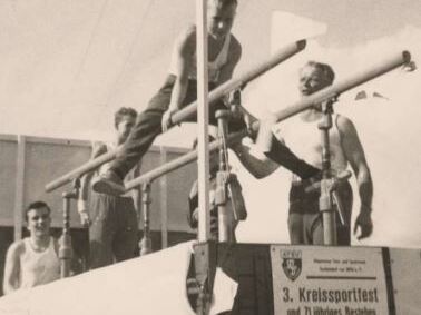 Gymnastik&uuml;bung an einem Reck mit mehreren M&auml;nnern, die an einem Wettkampf teilnehmen. Im Hintergrund ein Plakat.