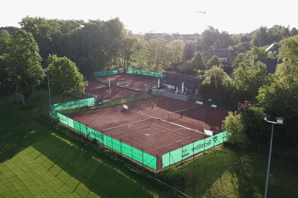 Luftaufnahme eines Tennisplatzes mit roter Erde, umgeben von Bäumen und einem Gebäude im Hintergrund.