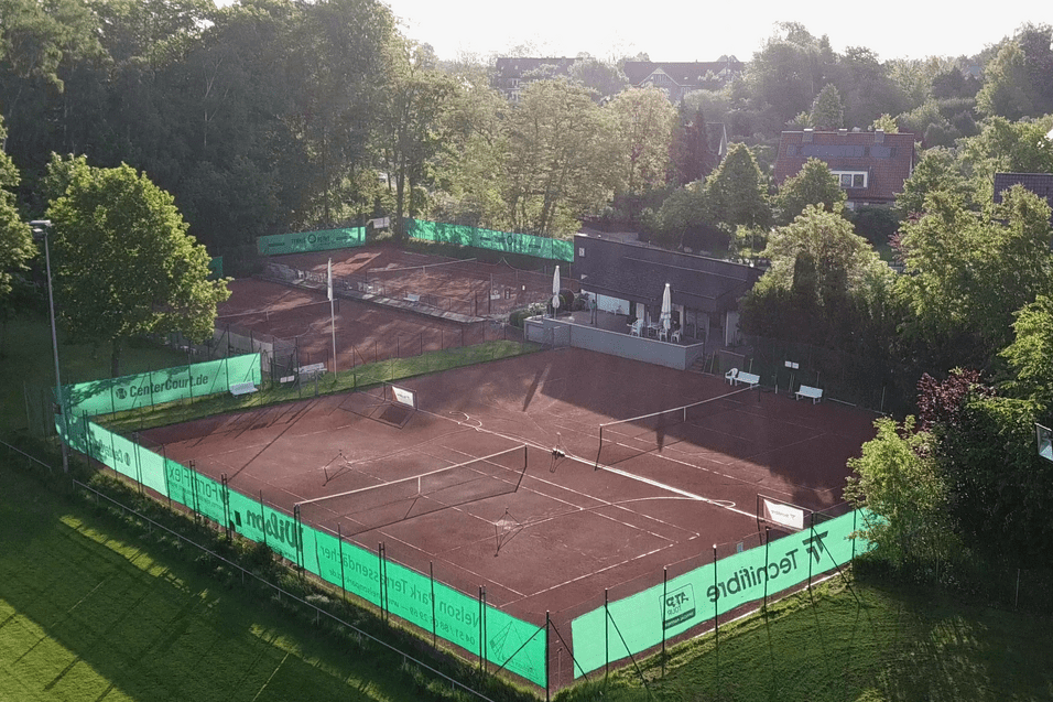 Luftaufnahme eines Tennisplatzes mit roter Erde, umgeben von B&auml;umen und einem Geb&auml;ude im Hintergrund.