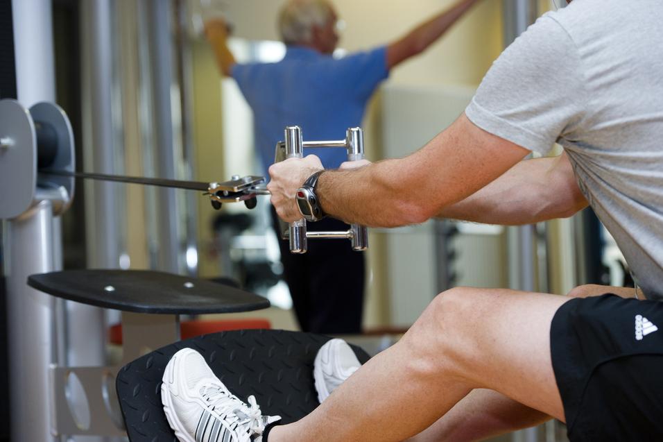 Mann trainiert im Fitnessstudio mit einem Kabelzuggerät, ein anderer Mensch im Hintergrund führt eine Übung aus.